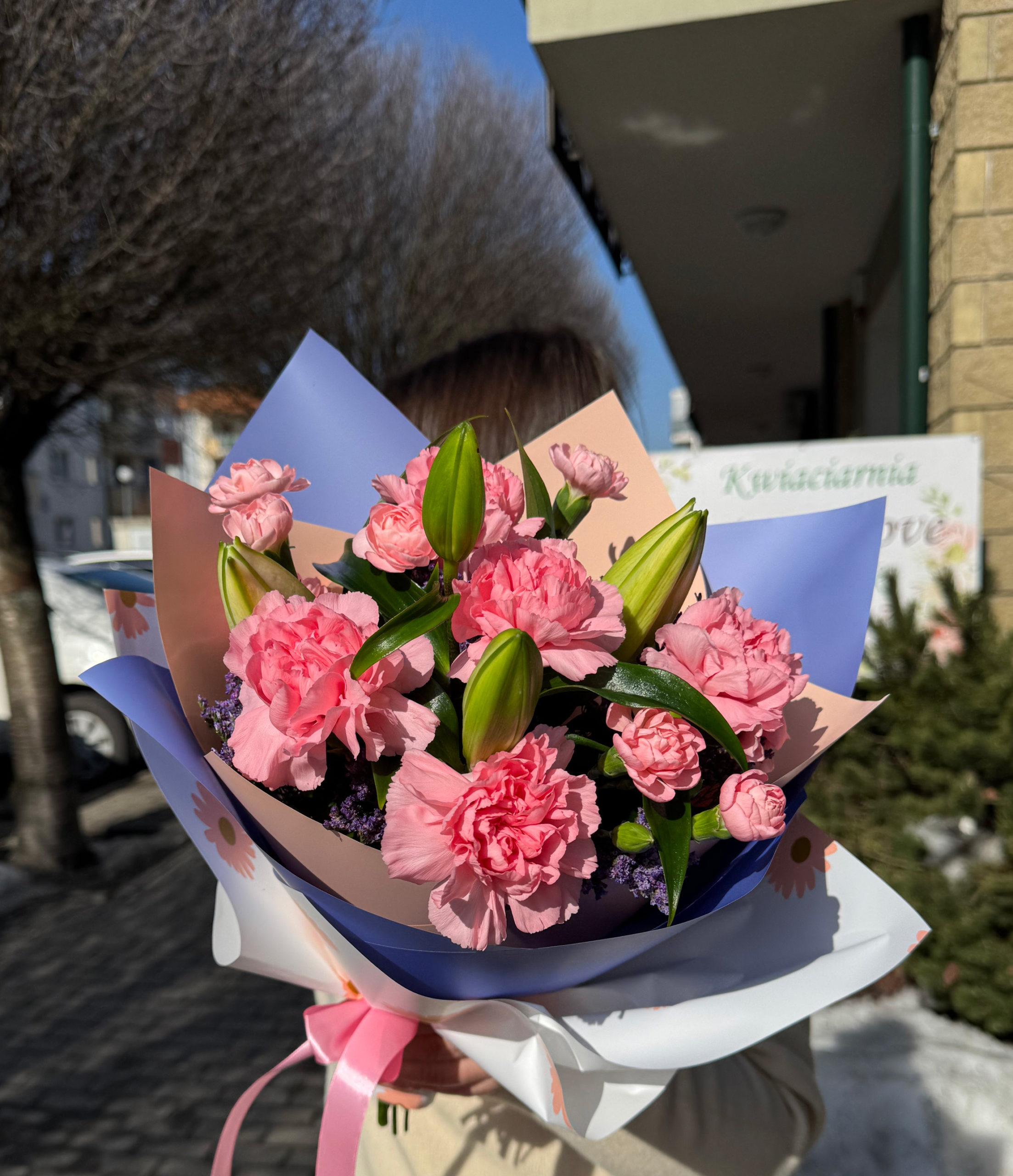 Biało różowo fioletowy bukiet z lilii, różowych goździków i limonium w papierze dekoracyjnym z kokardką